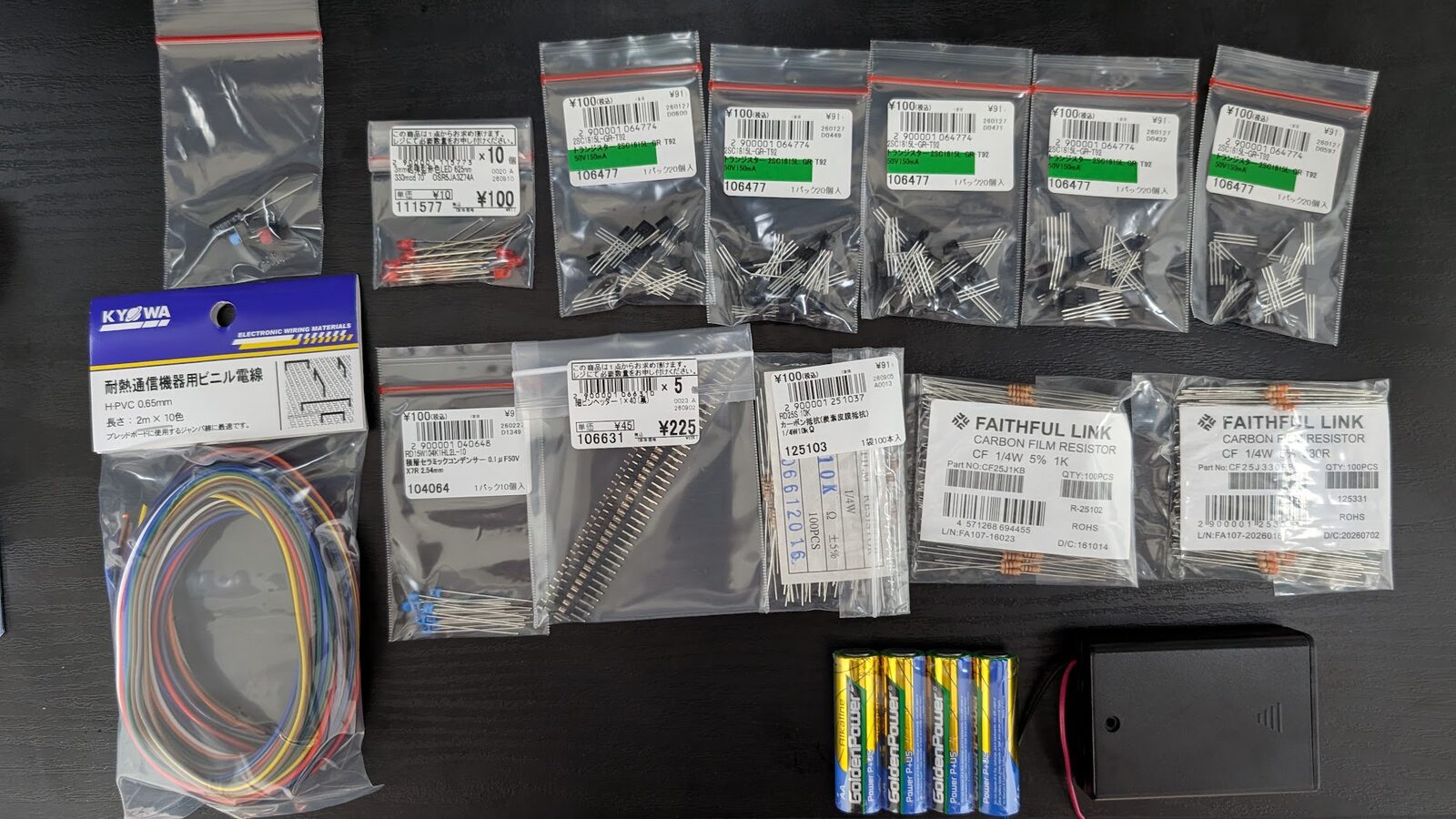 机に並べた回路部品。20 個入りのトランジスタの袋が 5 つ、カーボン抵抗が 3 袋、LED、積層セラミックコンデンサ、細ピンヘッダ、単 3 電池 4 本と電池ボックス、10 色のビニル電線が置いてある。